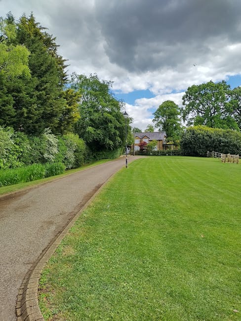 A neatly maintained garden scene featuring a curved gravel pathway bordered by a low brick edge, running alongside a expansive, well-manicured lawn with lush green grass. To the right, the lawn is sep