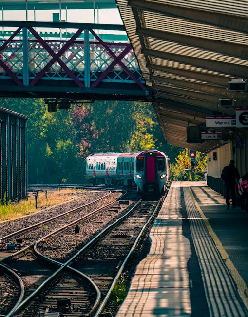 The image depicts a suburban train station platform with a modern train approaching in the distance. The platform is covered by a metallic canopy with horizontal slats, providing shade to waiting pass