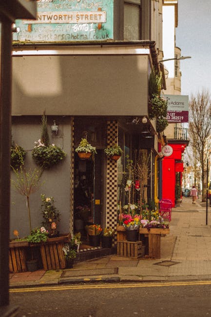 A display of various potted plants and flowers arranged on wooden shelves and tables outside a shop window, with large leafy green plants and smaller flowering pots including primroses in pastel-color